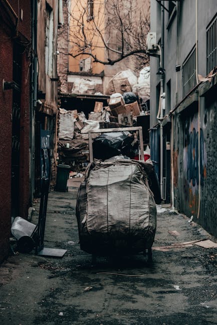 A municipal waste collection truck with a white exterior is parked on a narrow city street next to a row of older, weathered buildings with beige and gray facades. The truck's large rear loading compa