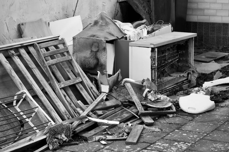 A collection of discarded household items and building debris piled on a paved outdoor area, including a wooden ladder with multiple rungs leaning against a wall, a damaged microwave with its front pa