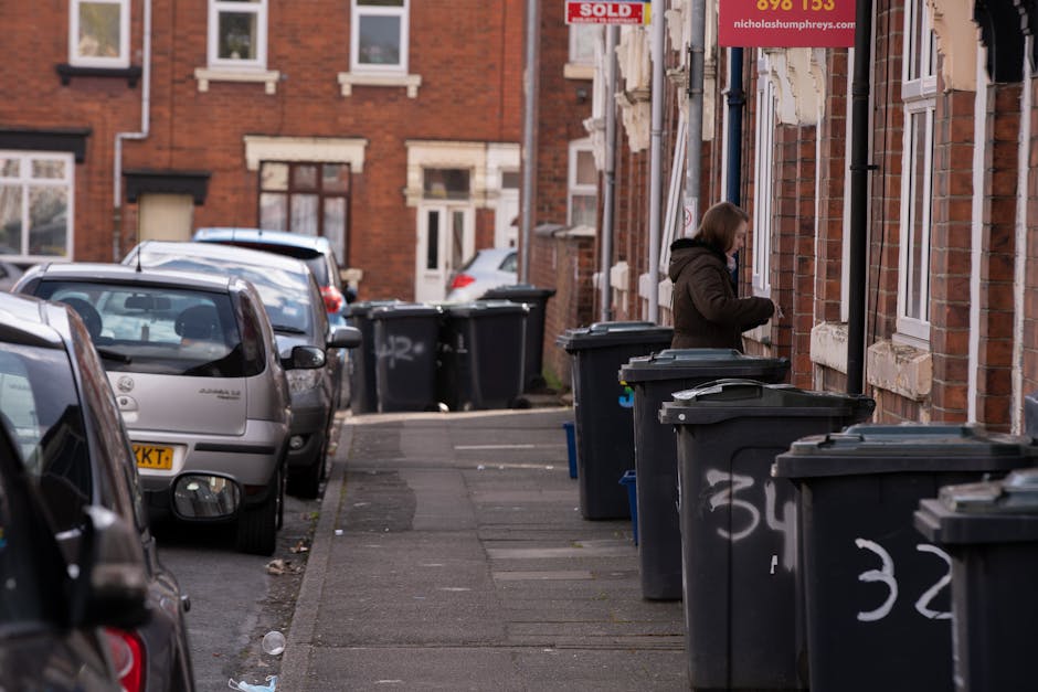 A row of outdoor wheelie bins lined up along a pavement next to a brick wall, featuring various colours including green, blue, red, and dark grey. The green bin in the foreground has a closed lid with