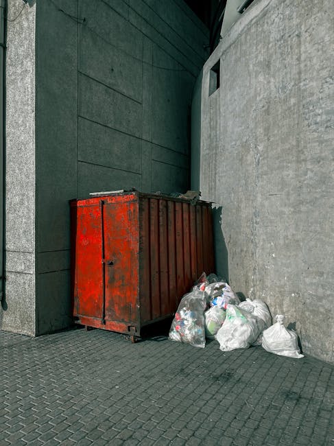 A white industrial refuse collection truck is parked on a narrow cobblestone street, with its rear open, revealing rusted and used interior components. A sanitation worker, dressed in a blue uniform a